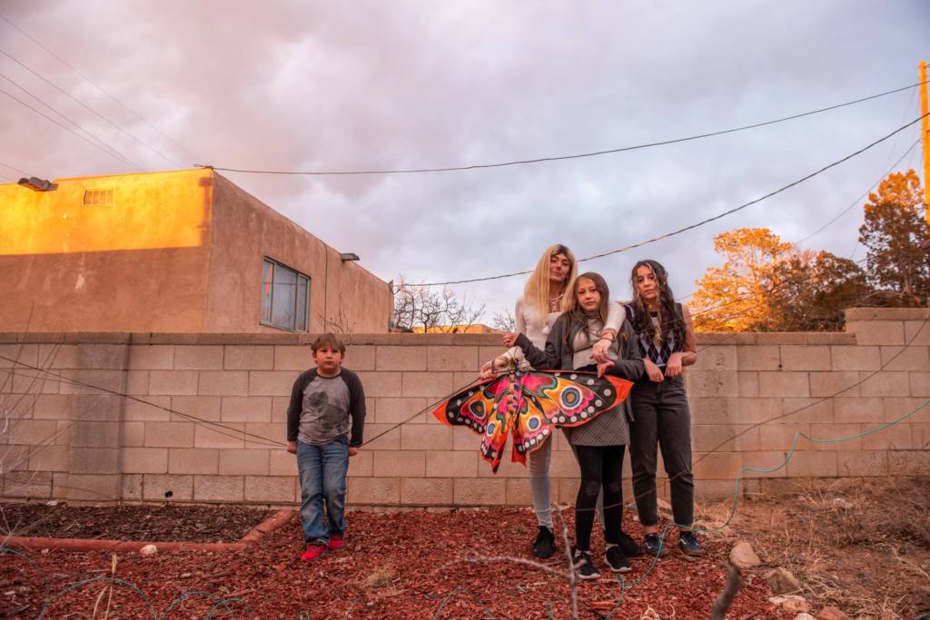 Elle (she/her) is pictured alongside her three young children in Santa Fe, New Mexico after celebrating themselves and their identities. Elle's hand is wrapped around her shorter daughter's shoulders, and they hold a large butterfly kite. To their left is Elle's son, and to their right, Elle's taller daughter. The sun is setting behind them.