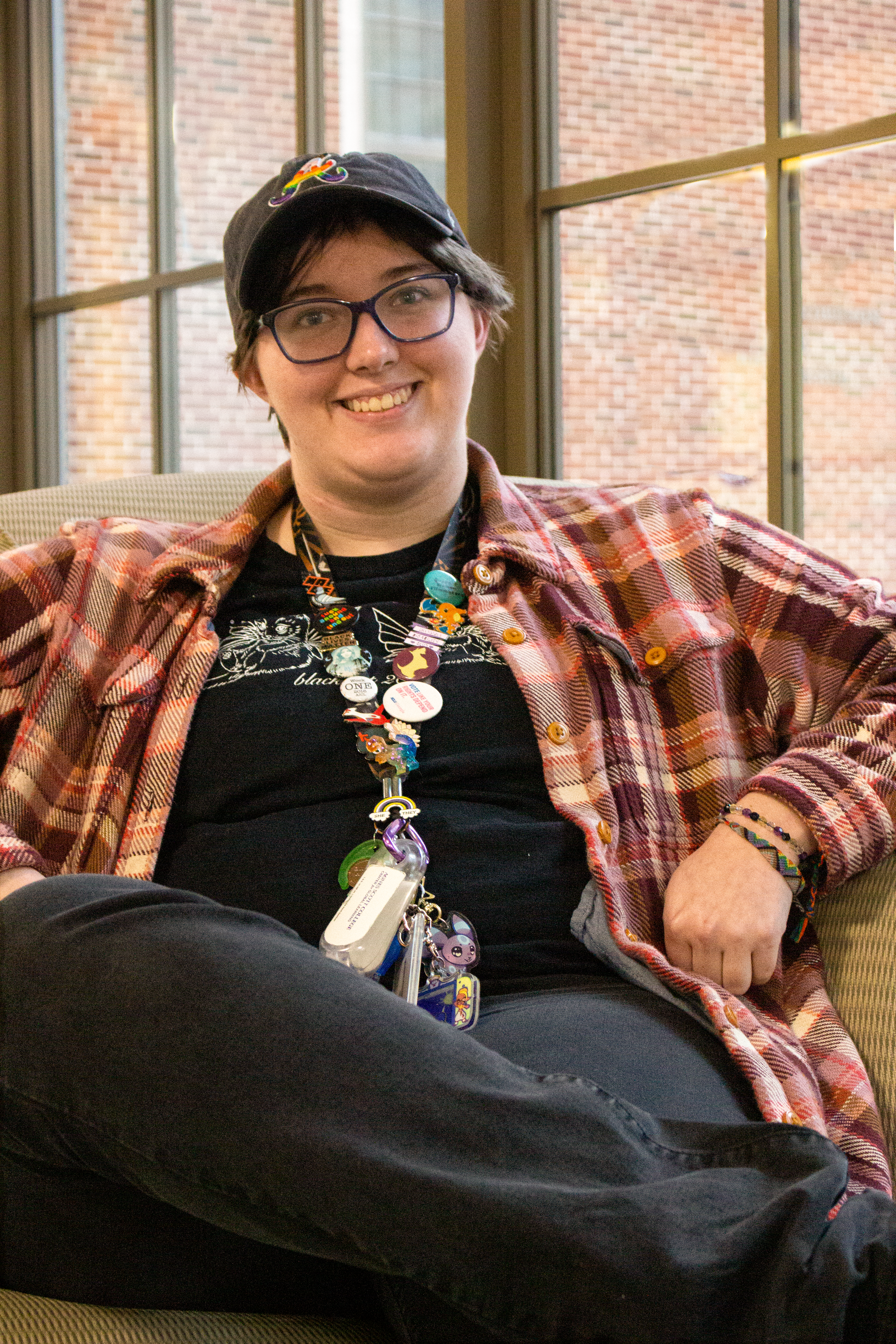 A person sitting cross-legged in a chair in front of a series of large windows. They are wearing black pants, a lanyard decorated with various pins, a black t-shirt, and a pink-and-brown flannel jacket.